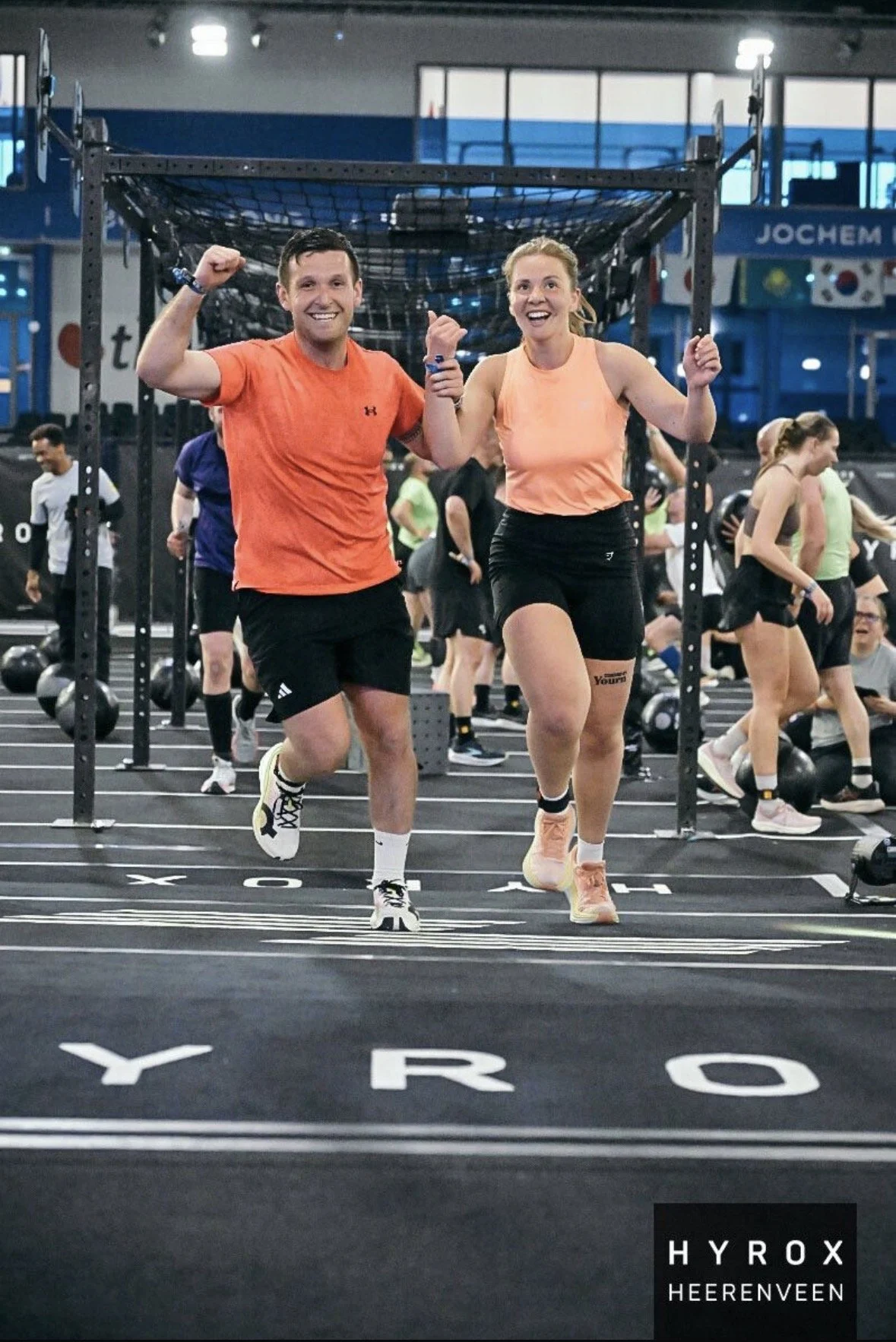 Twee mensen rennen samen op een atletiekbaan in een sportschool, terwijl ze elkaar de hand schudden en lachen. In de achtergrond zijn meerdere mensen te zien die aan krachttraining doen.