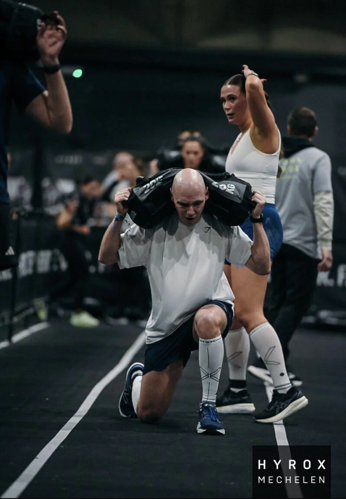 Een man knielt op de grond en tilt een gewicht op zijn schouders, terwijl een vrouw naast hem bezig is met haar haar. De achtergrond toont een sportschool met andere mensen.
