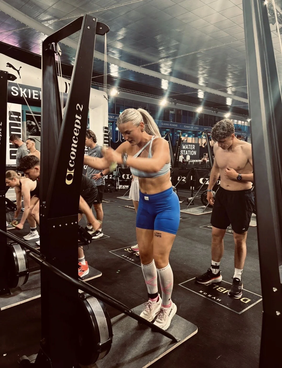 Mannen en vrouwen trainen in een fitnessruimte met verschillende apparaten, vrouwen dragen sportkleding en mannen trainen zonder shirt.