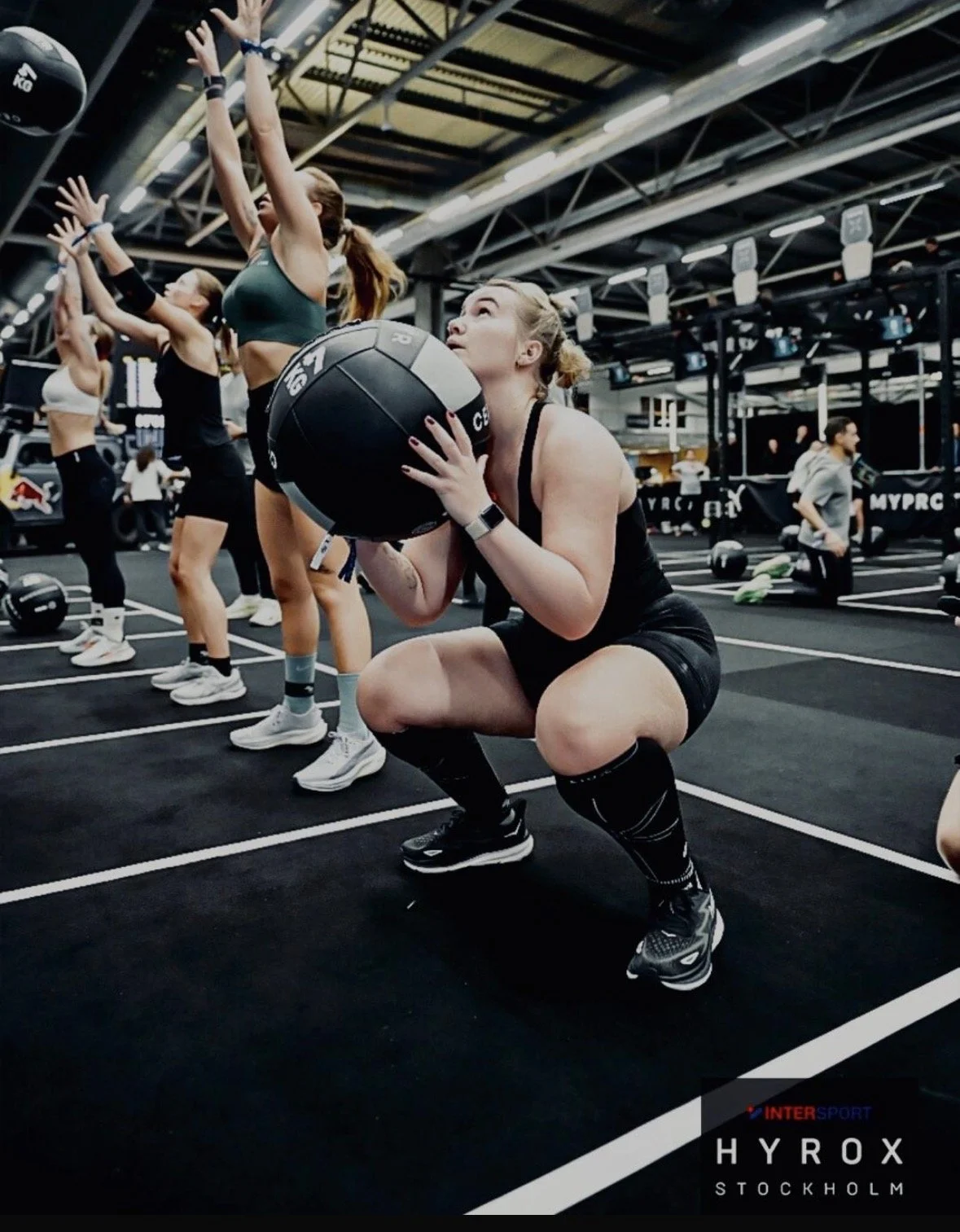 Vrouw die squats doet met een medicijnbal in een fitnessruimte, omringd door anderen die oefeningen uitvoeren.