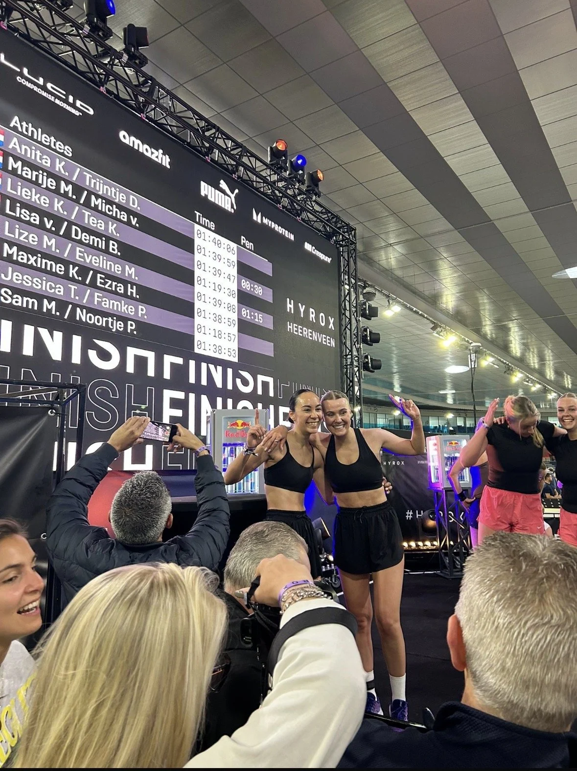 Twee vrouwen in sportkleding vieren op het podium bij een sportwedstrijd, met een digitale broadcast achtergrond en anderen in de menigte die foto's maken.