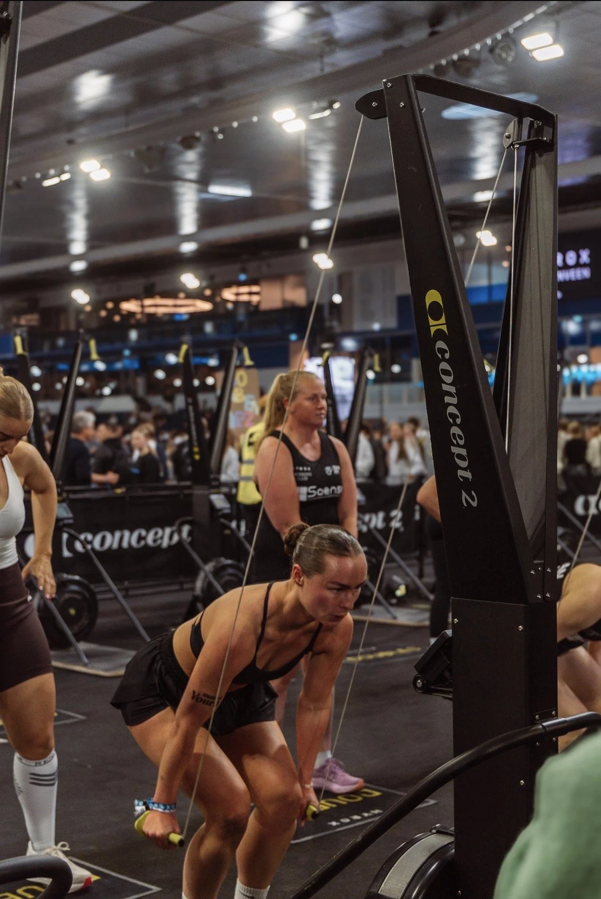 Vrouw die roeitraining doet in een sportschool, omringd door andere vrouwen en professionele roeitrainers.