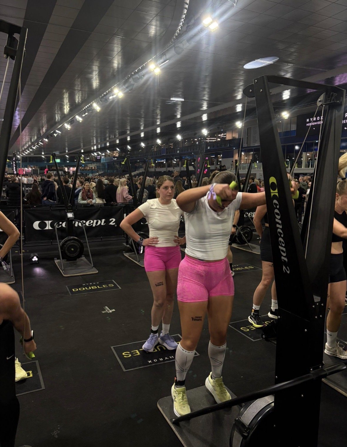 Binnenbeeld van een druk evenement met vrouwen die fitnessoefeningen doen op een sportschoolvloer, onder kunstlicht.