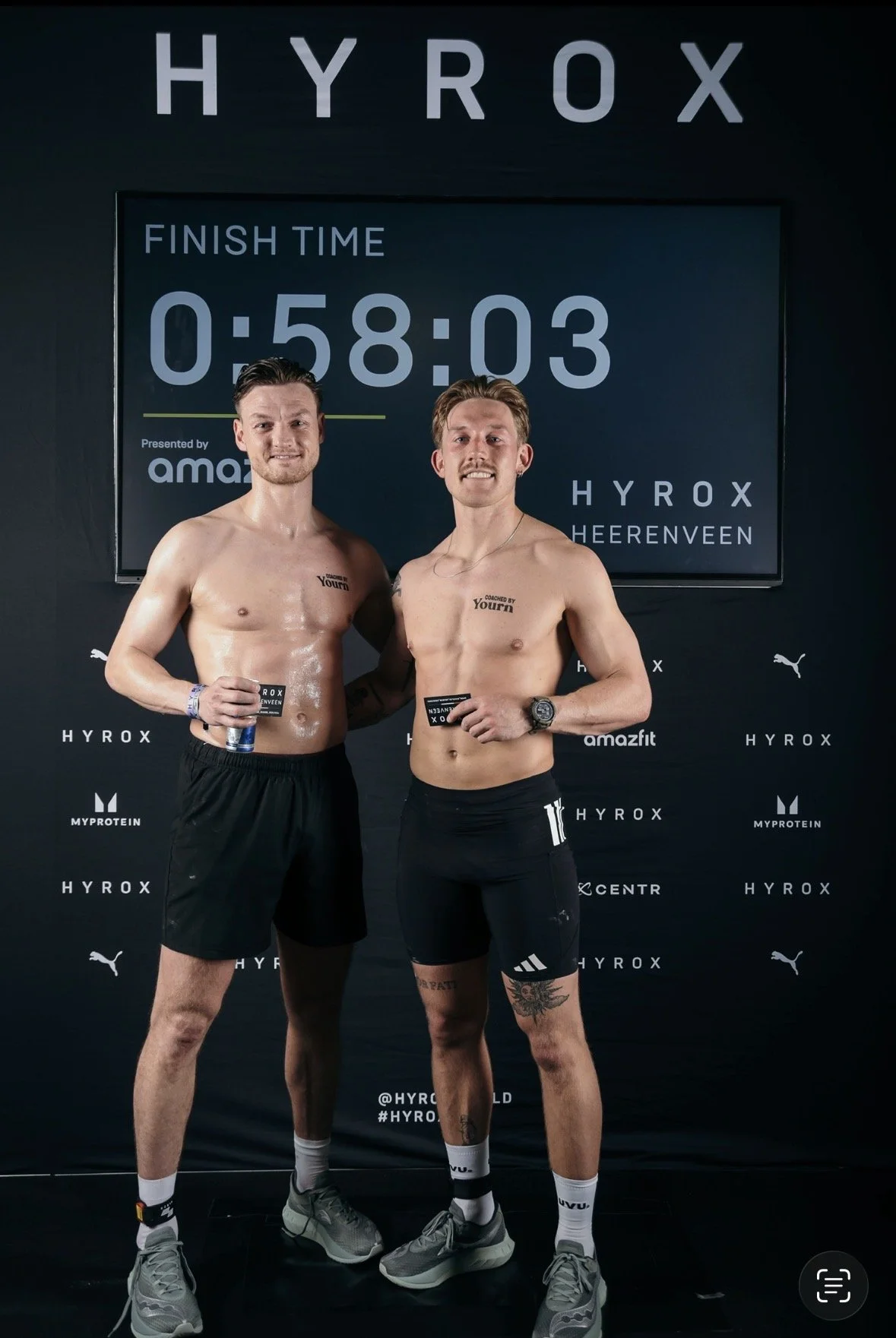 Twee shirtsloze mannen na een sportevenement, met een finish tijd op een scherm achter hen van 58 minuten en 3 seconden, bij Hyrox in Heerenveen, Nederland.