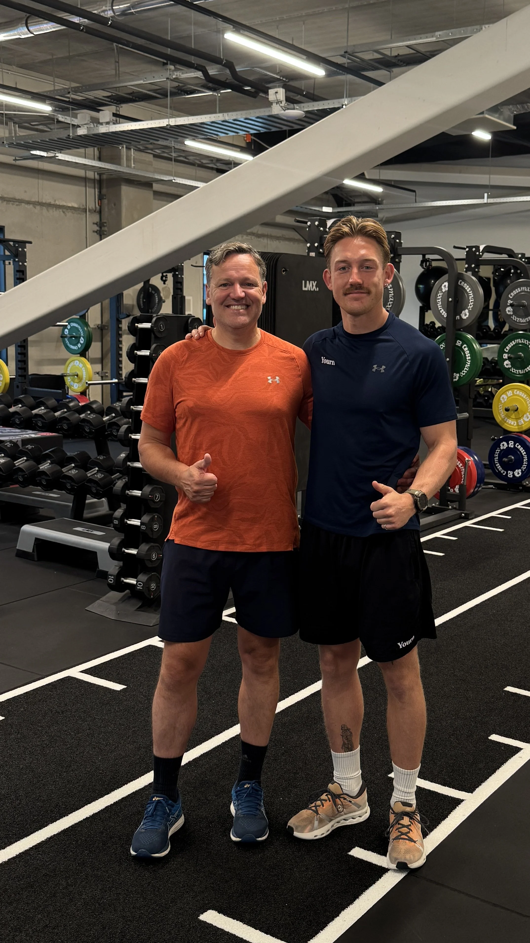 Twee mannen in sportkleding staan naast elkaar in een sportschool, ze glimlachen en maken een vuistgebaar. Achter hen bevinden zich gewichten en fitnessapparatuur.