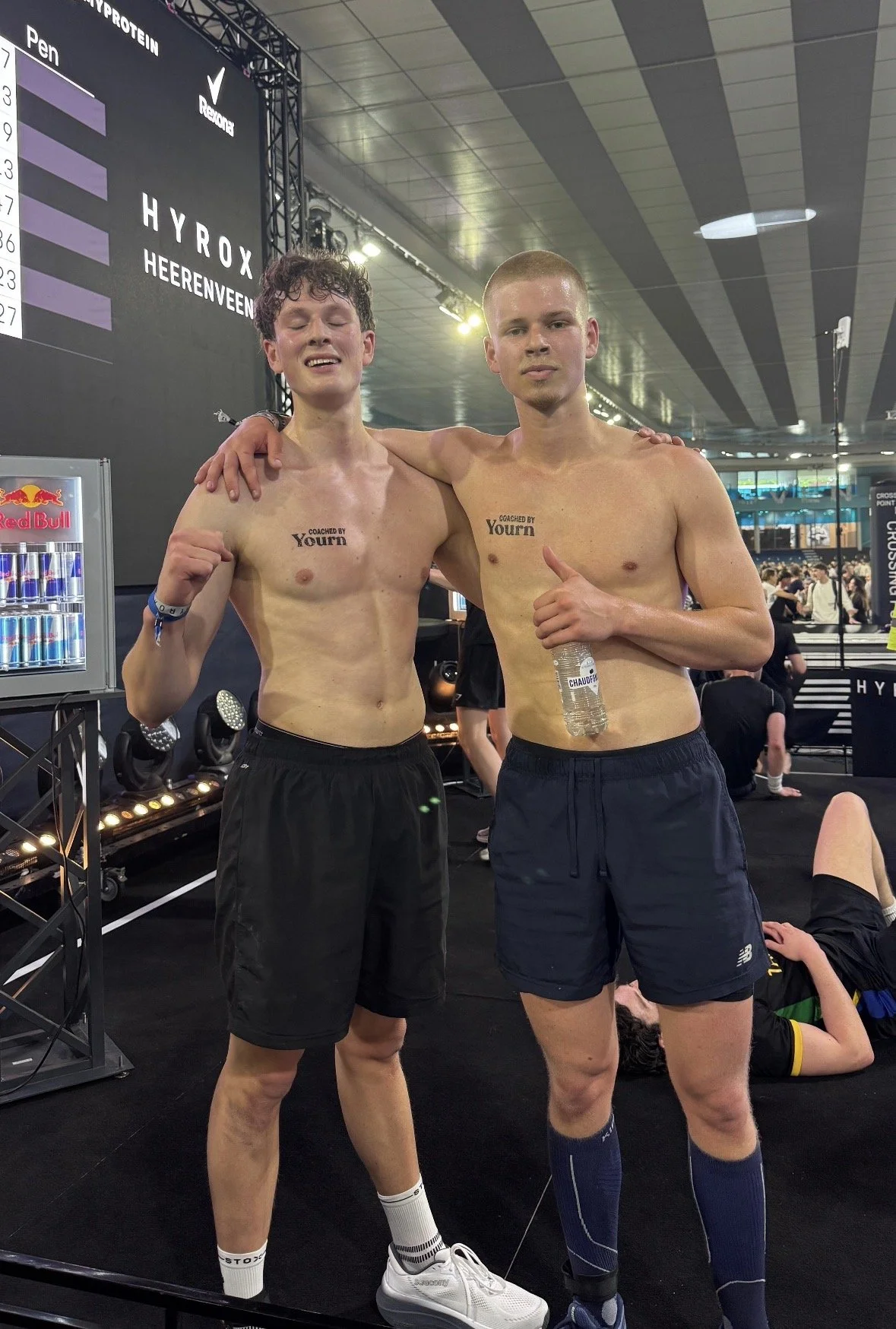 Twee jonge mannen zonder shirts staan naast elkaar in een sporthal, ze lachen en maken schouderkus en gebaren. Ze dragen sportkleding en hebben tatoeages die zeggen 'Coached by Yourn'.