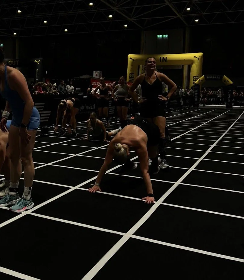 Vrouwen doen een fitnessoefening op een sportbaan, met een overkapping en een geel startblok in de achtergrond.