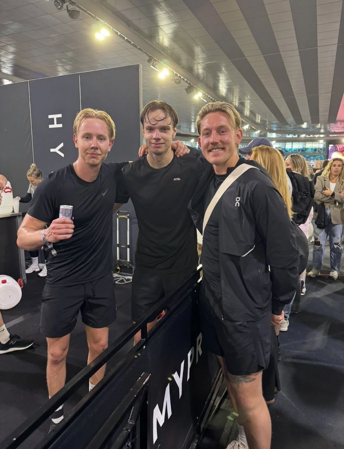 Drei jonge mannen in sportkleding staan samen en glimlachen voor de camera in een drukke hal of sportevenement. Eén man houdt een flesje water.