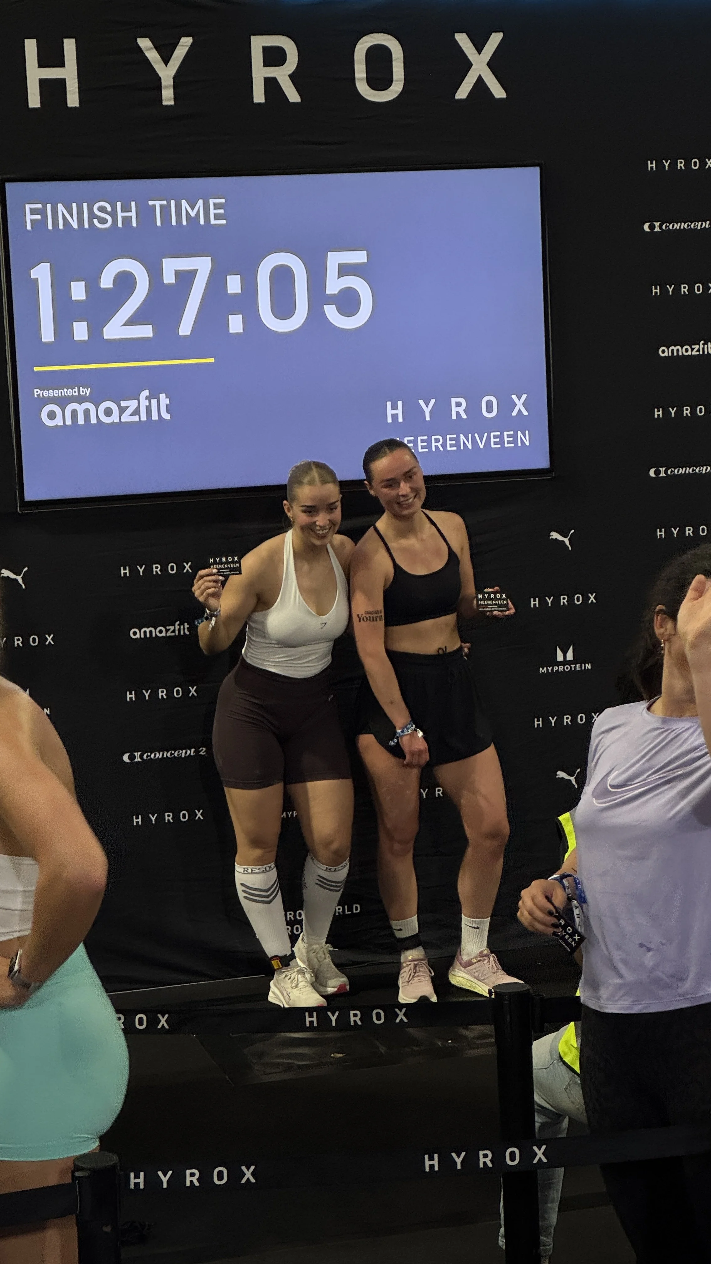 Twee vrouwen staan op het podium bij het einde van een hardloopwedstrijd, ze tonen hun medaille en glimlachen. Op de achtergrond is een groot scherm met de eindtijd van 1:27:05 en de tekst 'Finish Time'.