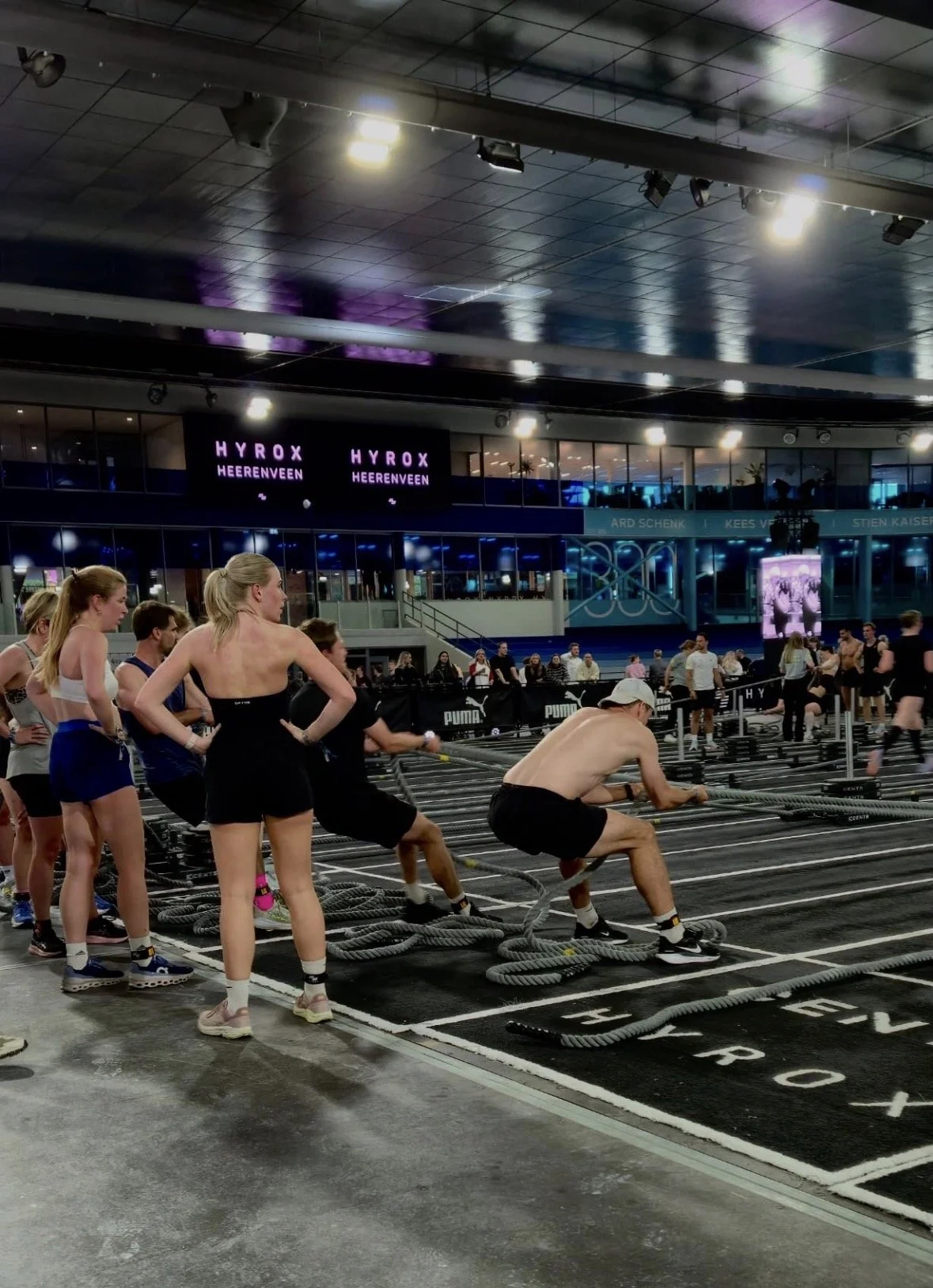 Mensen die aan krachttraining doen met sleeptouwen in een sporthal, met digitale schermen en een publiek op de achtergrond.
