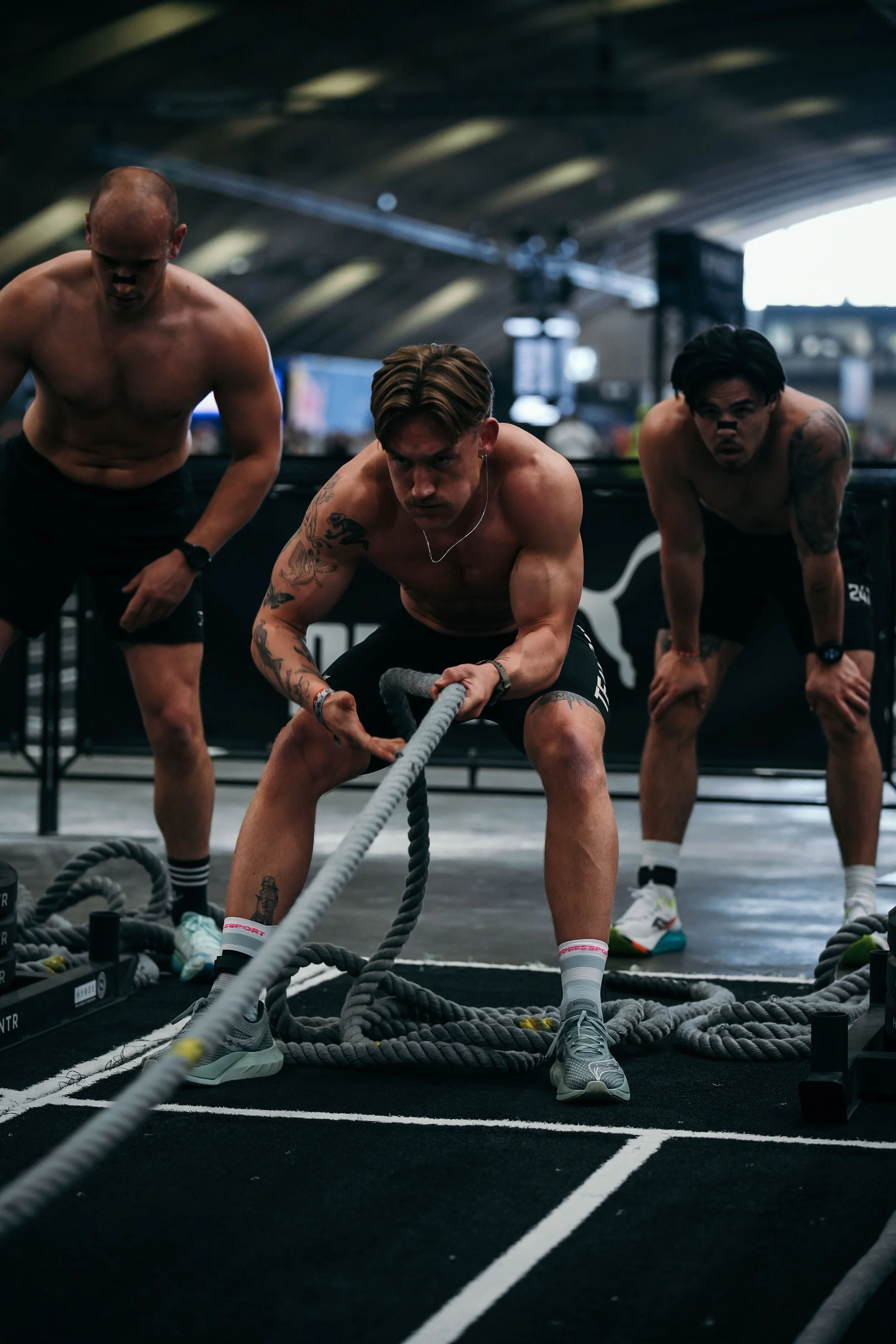 Drie shirtloze mannen bezig met een workout op een indoor sportschool met touwen en fitnessapparatuur.