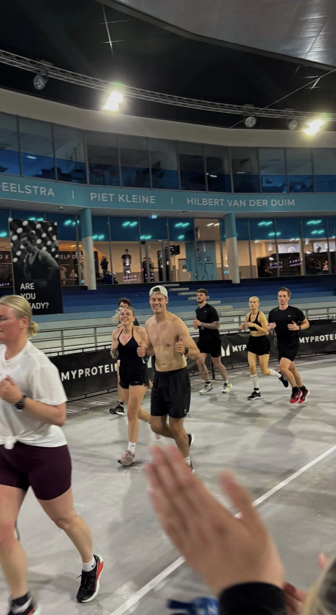 Mensen die hardlopen in een binnenbaan stadion, met een banner van Myprotein en de namen van de straten op de achtergrond.