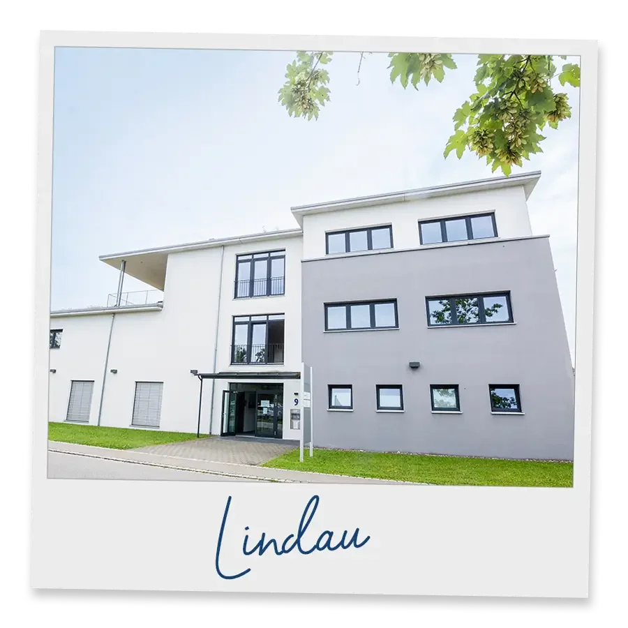 Mehrstöckiges modernes Wohngebäude mit weißen und grauen Fassaden, mehreren rechteckigen Fenstern und einer Eingangstür, umgeben von Grünflächen und einigen Baumzweigen im Vordergrund.