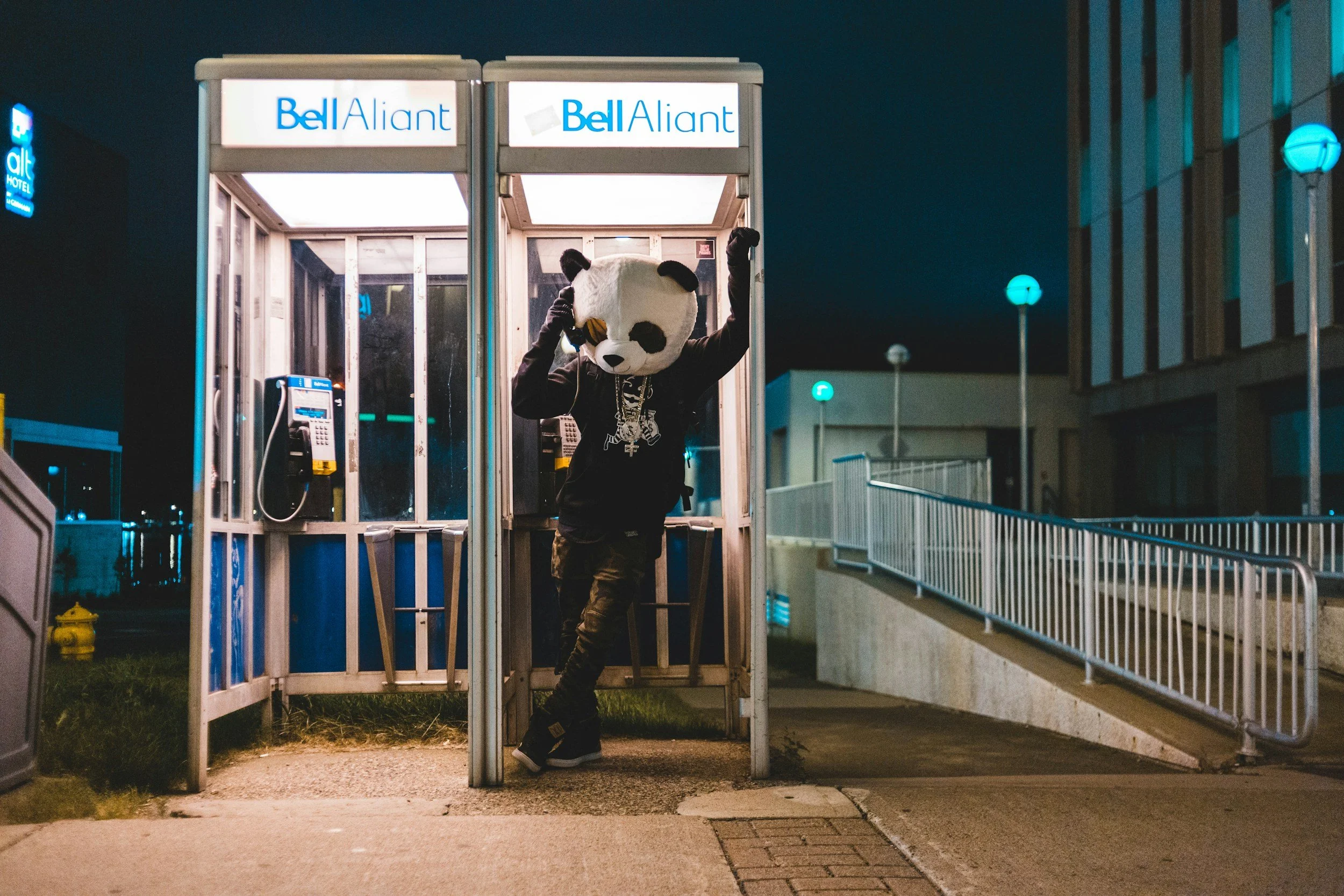 A person wearing a panda mask and black clothing, standing in a phone booth at night, adjusting the mask.