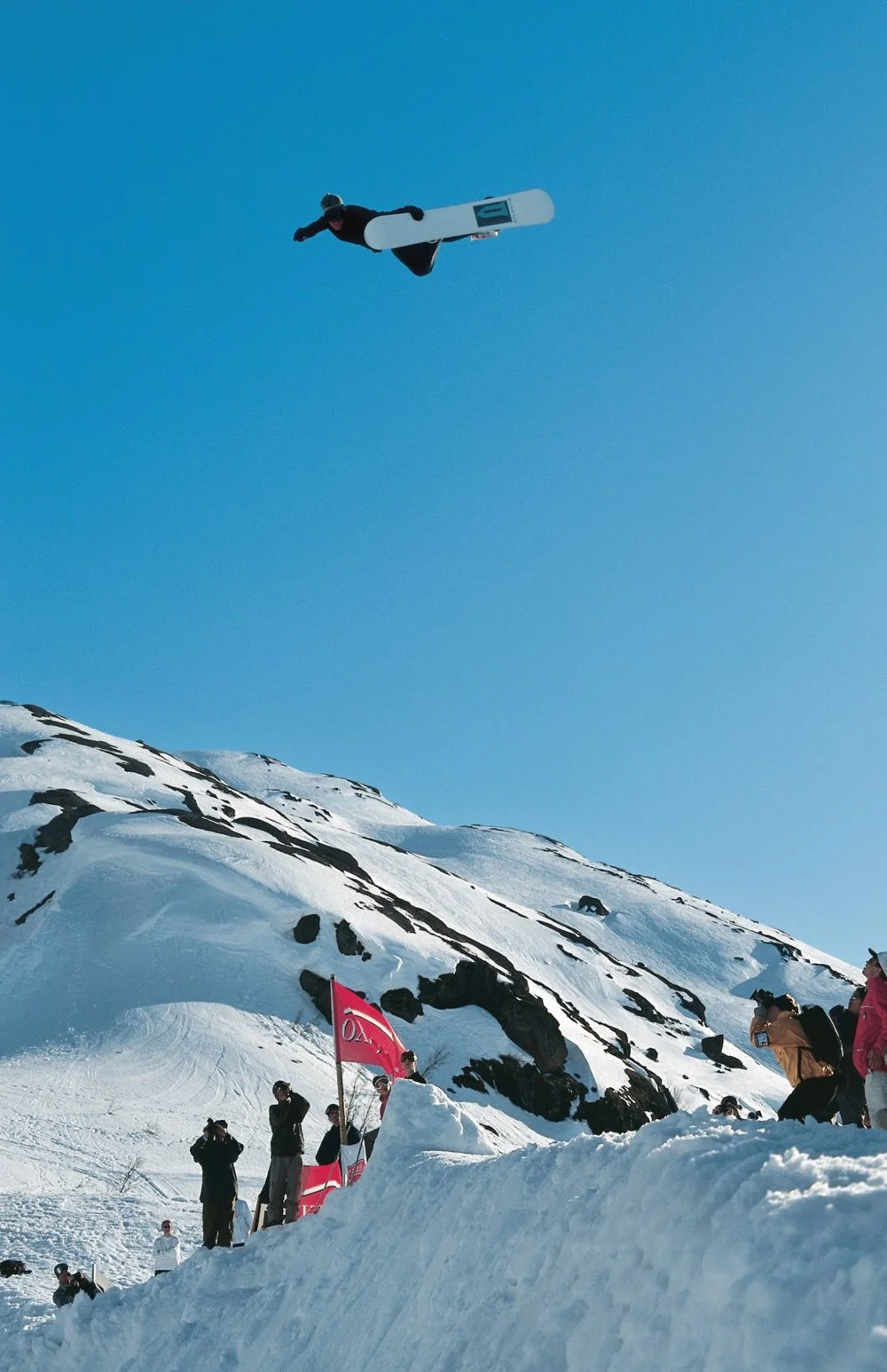 A snowboarder in mid-air performing a jump over a snowy slope during a snowboarding event with spectators and flags below.