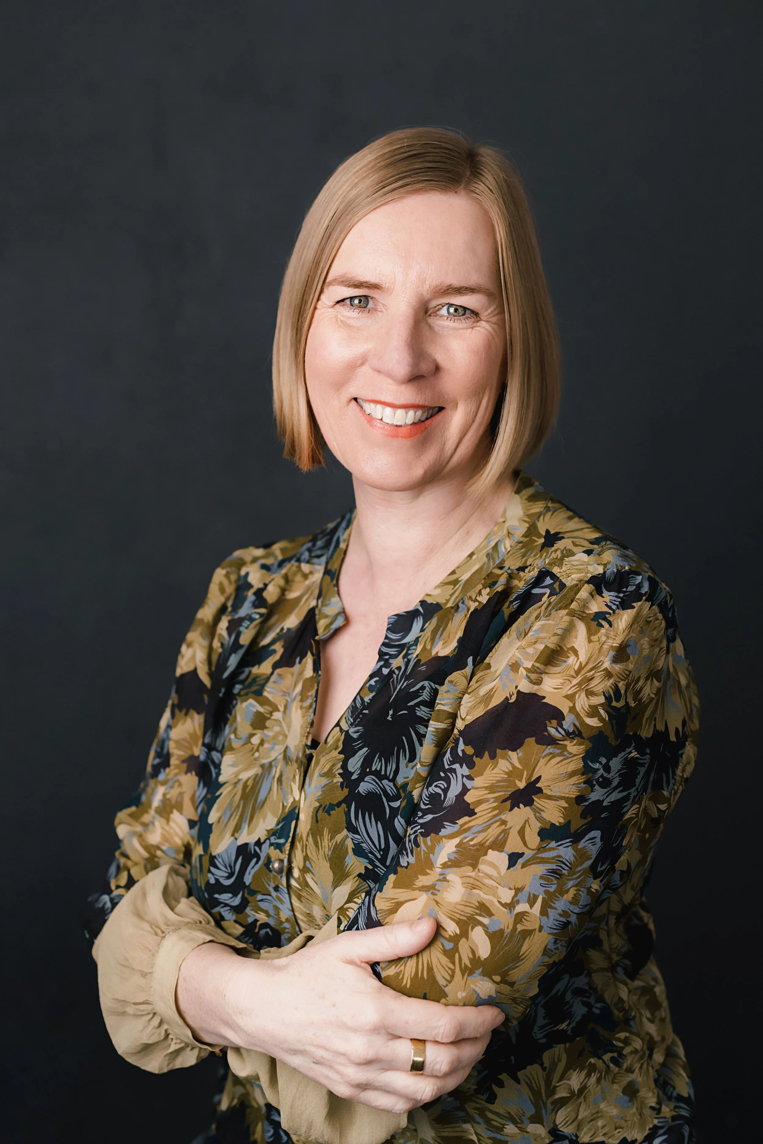 A woman with shoulder-length blonde hair, wearing a floral-patterned blouse in shades of black, gold, and beige, smiling with crossed arms against a dark background.