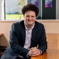 A man with curly hair wearing a dark blazer and white shirt, seated at a wooden table in an office or classroom setting.