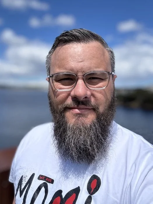 A man with glasses and a beard standing outdoors near a body of water on a sunny day with a blue sky and clouds.