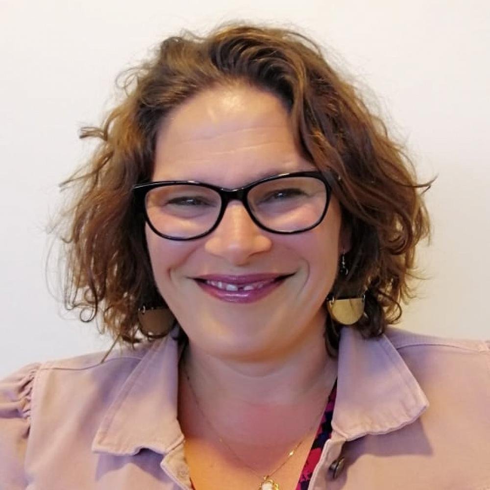 A woman with curly brown hair, glasses, and earrings smiling in front of a plain white background.