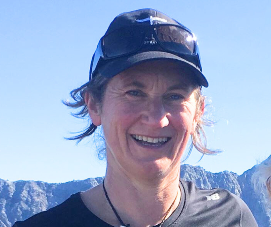 A woman outdoors smiling, wearing a cap, sunglasses, and athletic clothing, with mountains in the background.