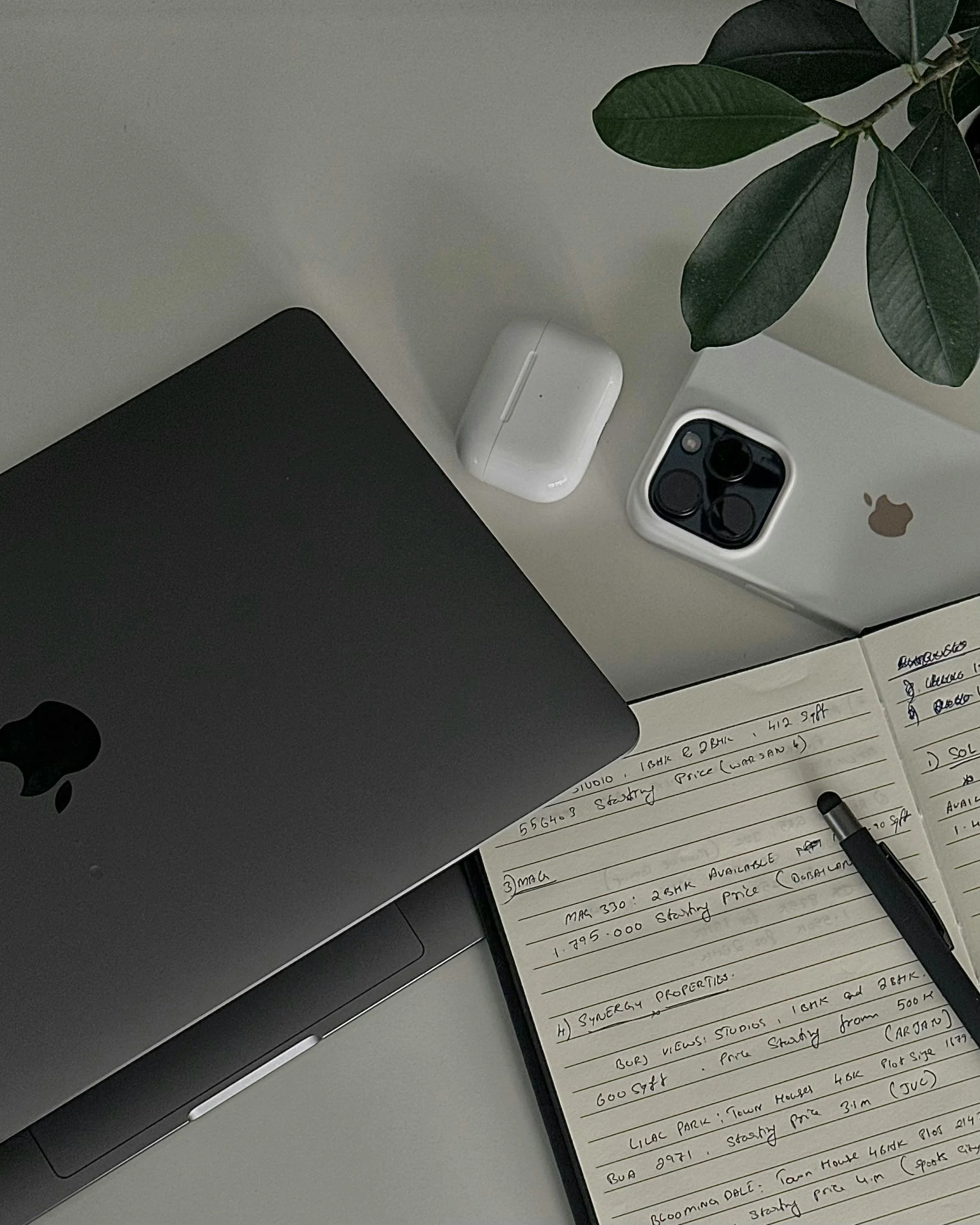 Flat lay of a workspace with a closed computer, wireless earbuds case, smartphone, notebook with handwritten notes, and a pen, next to a potted plant.