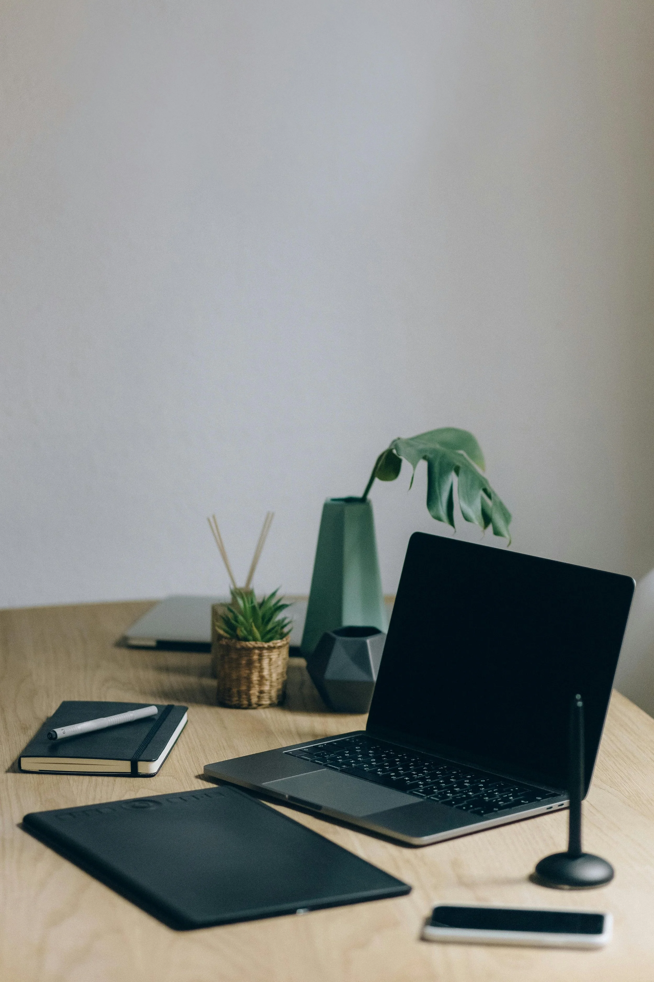 Minimalist workspace with a laptop, a drawing tablet, a smartphone, a closed notebook with a pen, and decorative plants on a wooden desk.