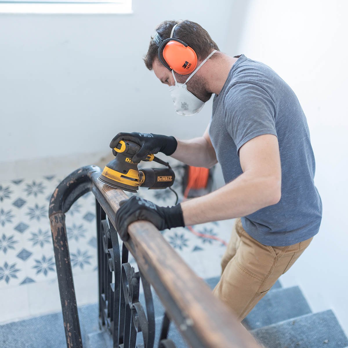 A man wearing safety ear protection, a face mask, and gloves is sanding a wooden handrail with a yellow and black DeWalt electric sander on a staircase.