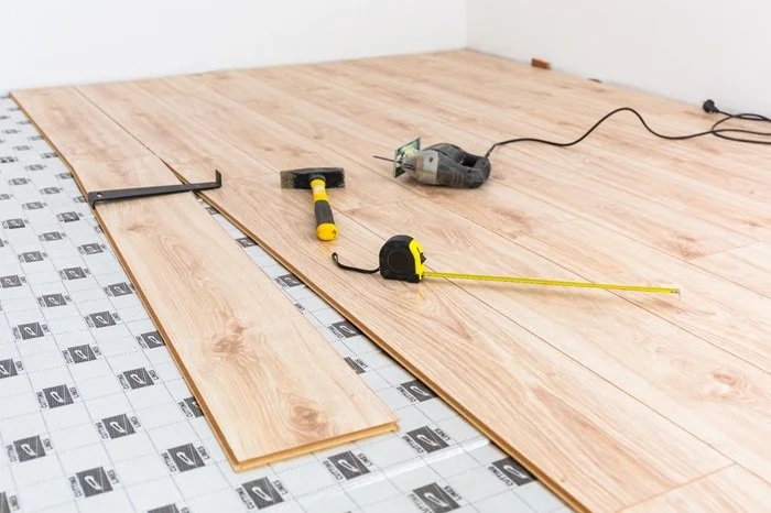 Wood flooring installation tools, including a hammer, tape measure, electric saw, and T-square, on a partially installed wooden floor with underlayment underneath.