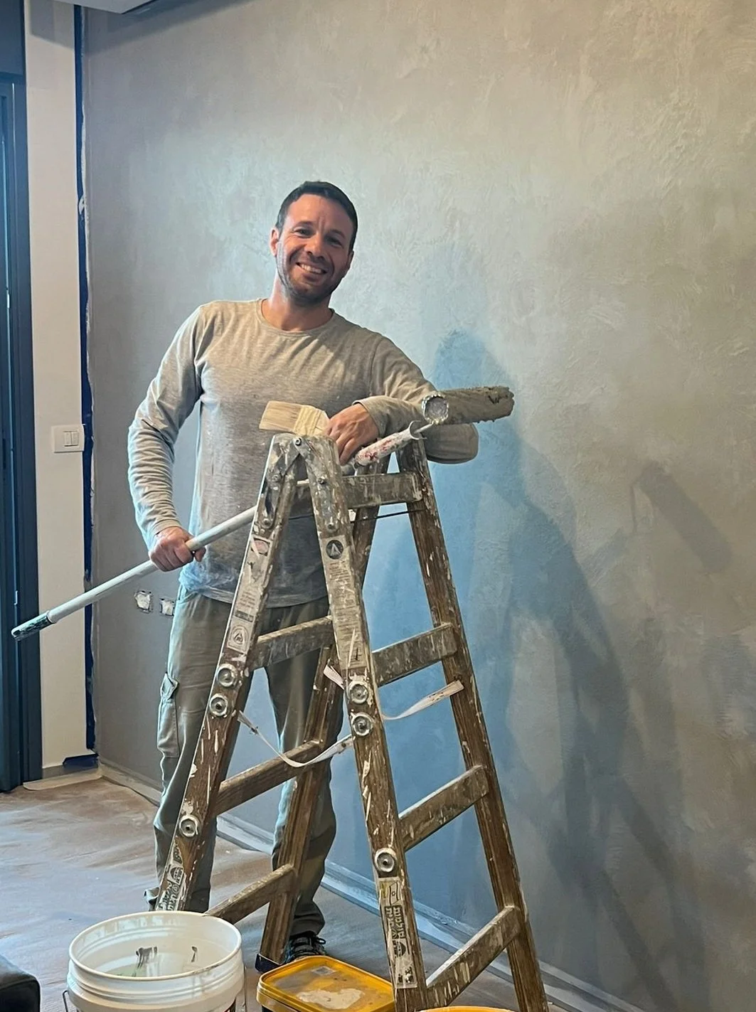A man smiling while painting a wall in a room, standing on a wooden ladder, with painting supplies nearby.