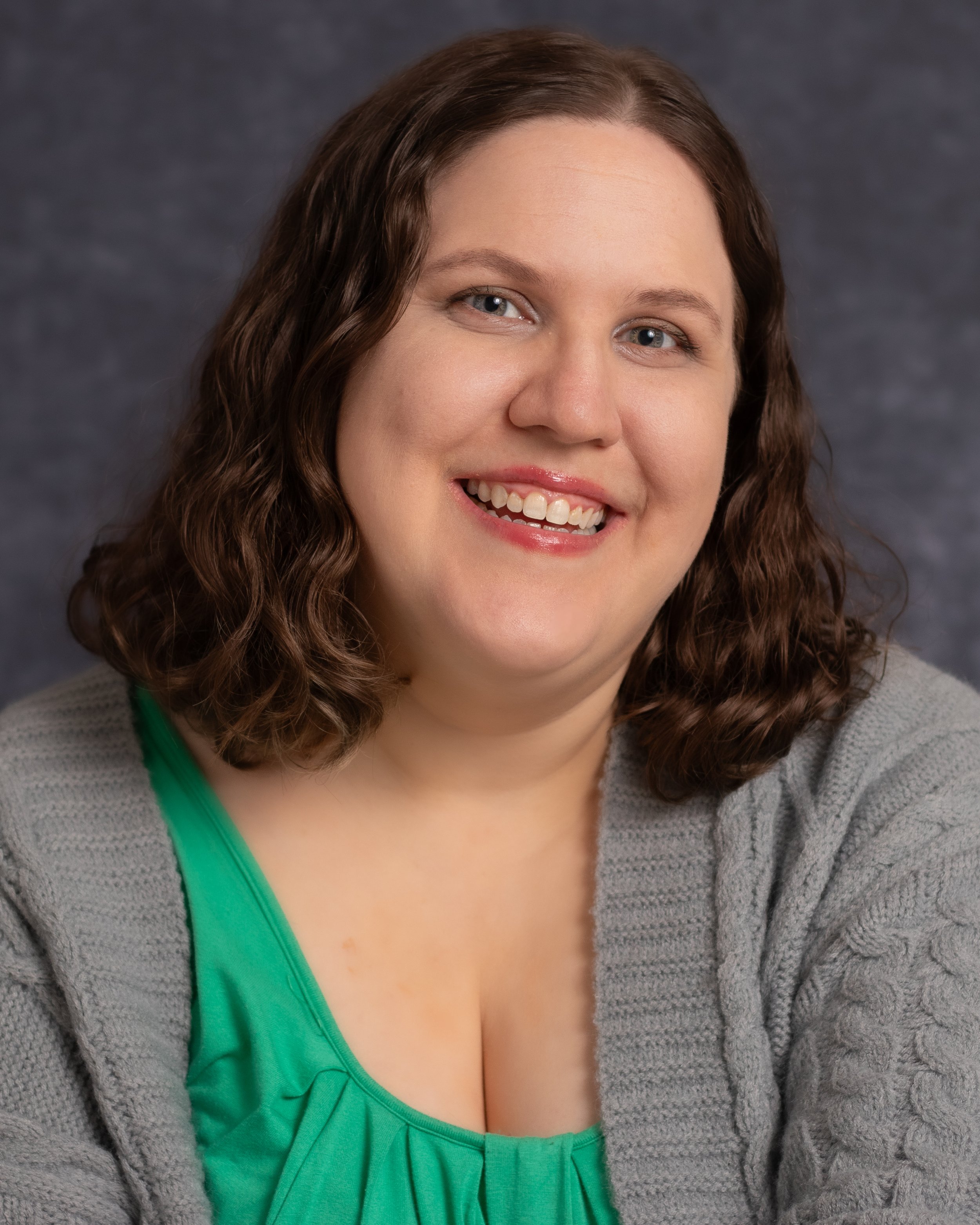 A smiling woman with shoulder-length curly brown hair, wearing a green top and a gray cardigan, posing against a dark gray background.