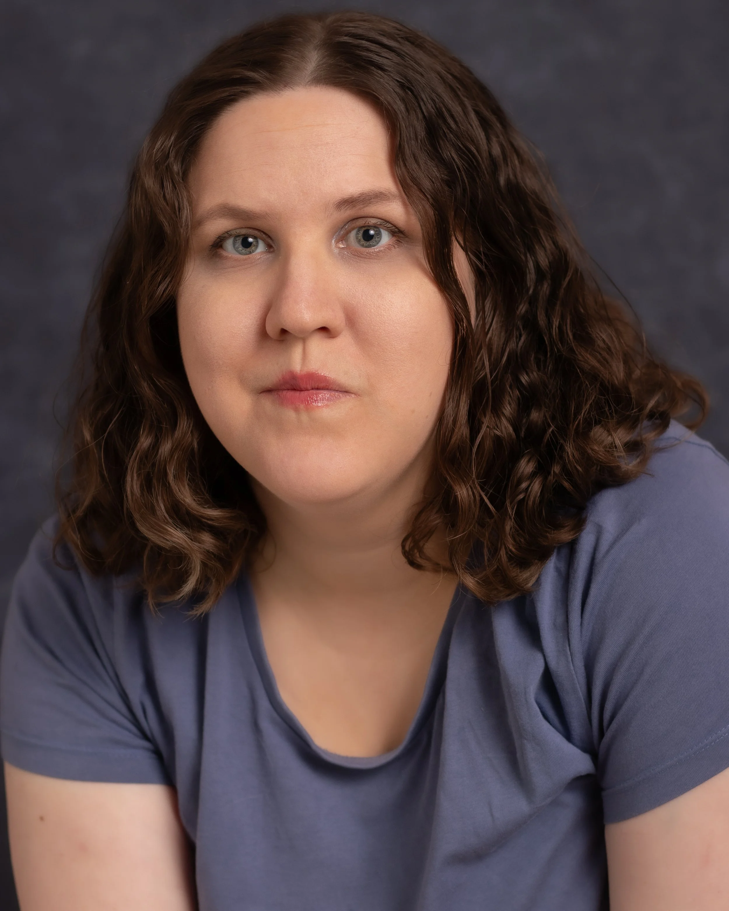 A woman with shoulder-length brown wavy hair and blue eyes, wearing a blue t-shirt, looking directly at the camera against a dark background.