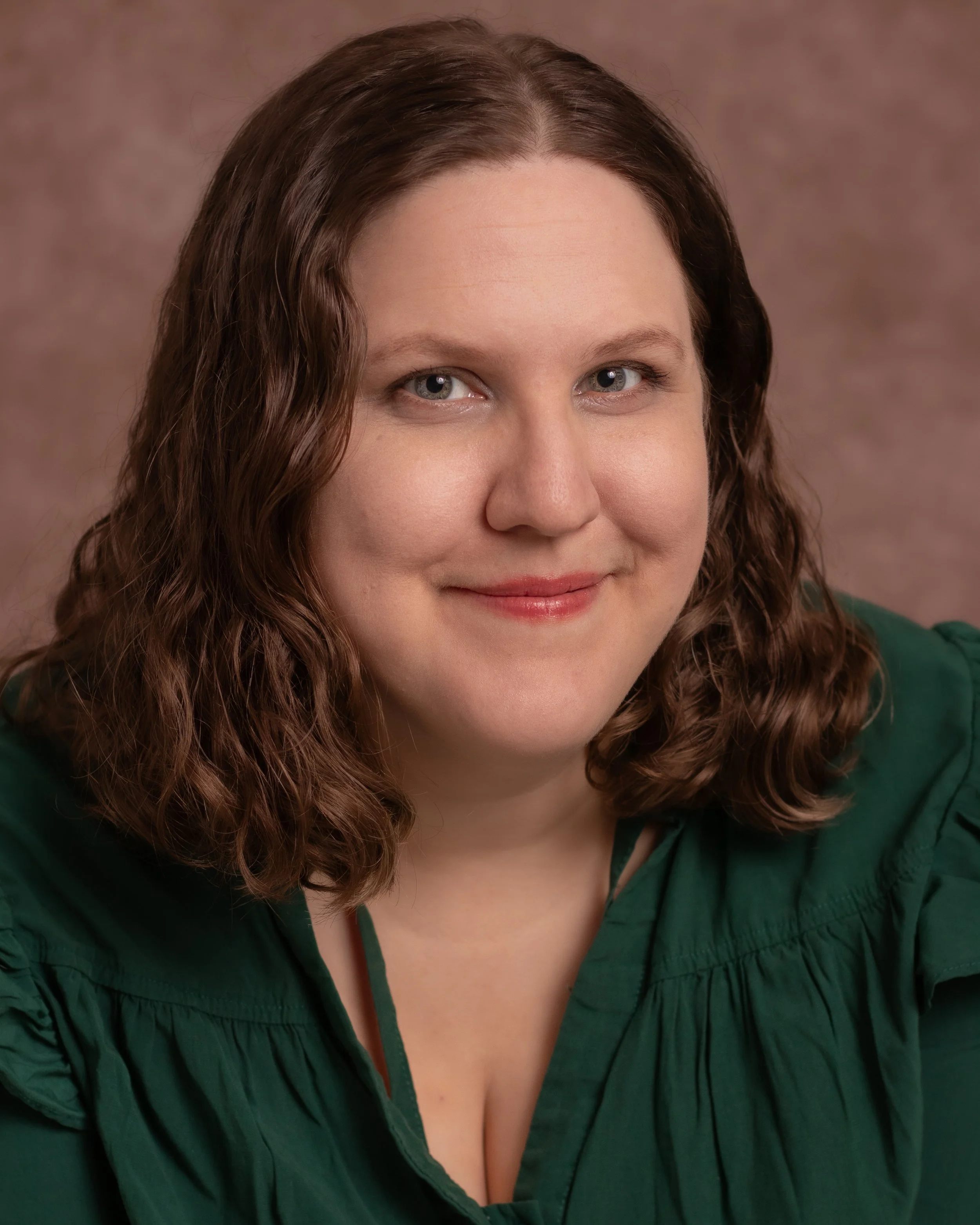 Portrait of a woman with shoulder-length brown curly hair, smiling, wearing a dark green blouse, against a brown background.