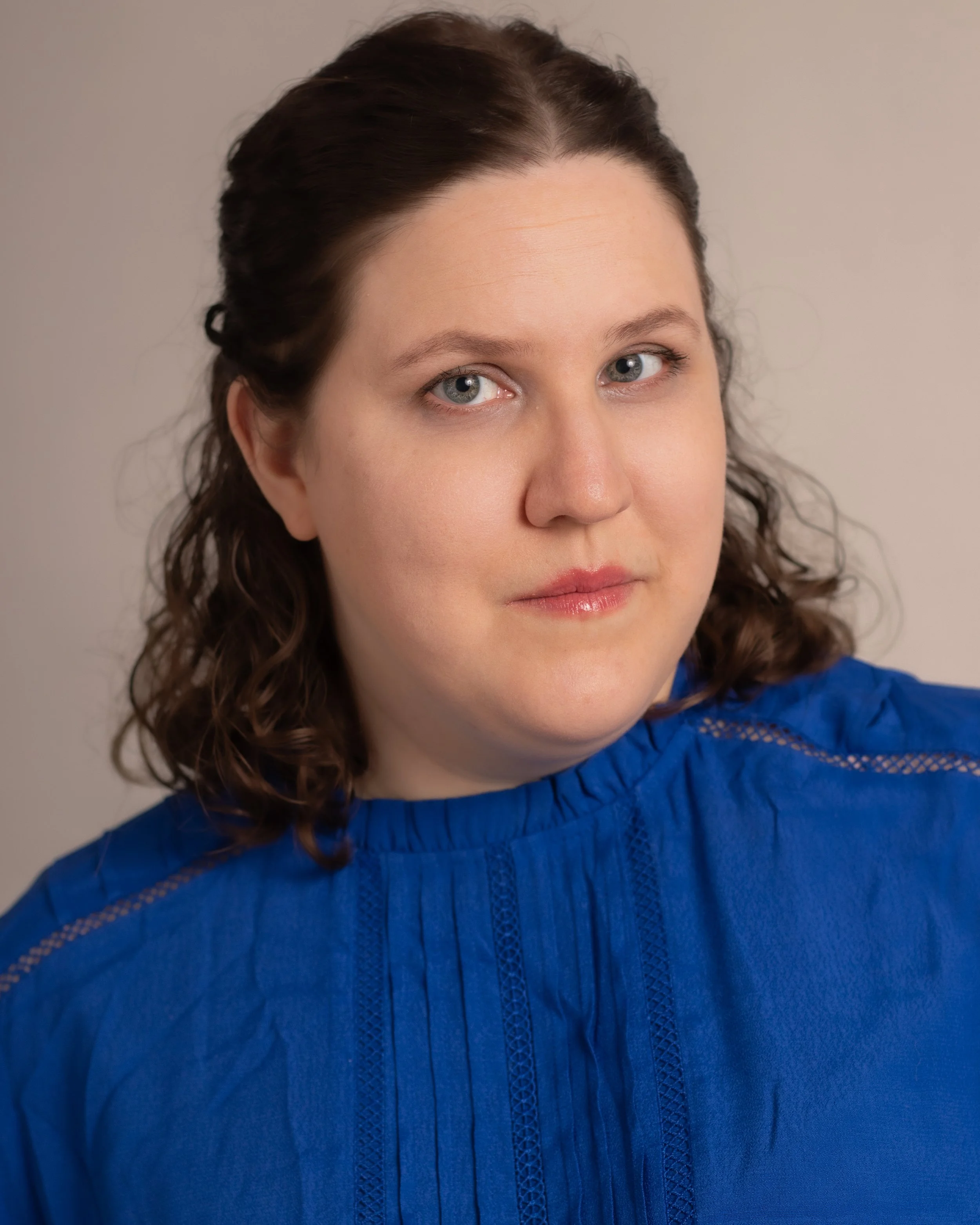 A woman with shoulder-length curly brown hair, light skin, and blue eyes, wearing a blue blouse, looking at the camera with a neutral expression.