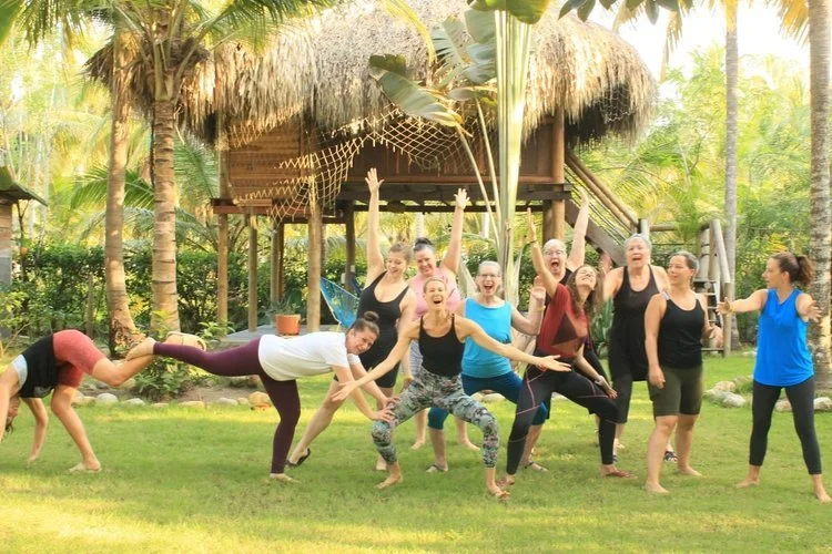 A group of women happily posing and having fun outdoors in front of a tropical hut, some making funny poses and others smiling and cheering.