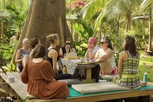 Group of women sitting around a table outdoors with lush green trees and plants in the background, enjoying a meal or conversation.