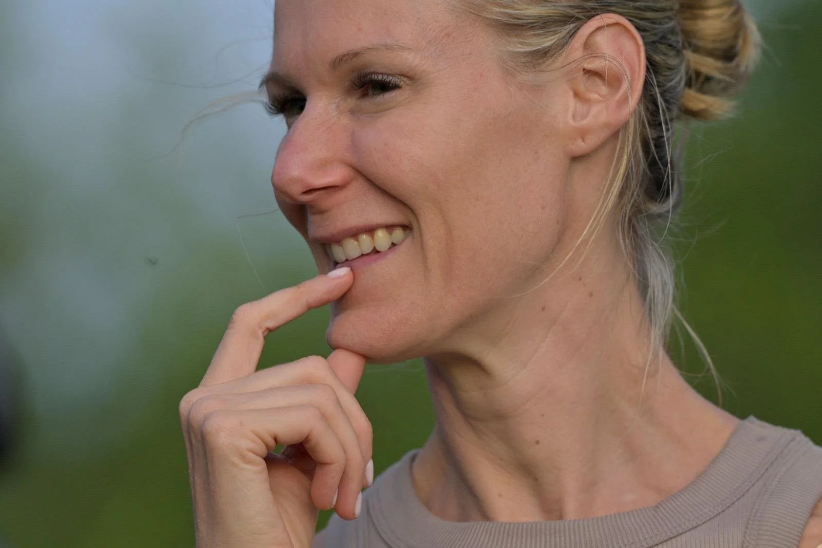 A close-up portrait of Eva Still smiling with her finger on her lips, set against a blurred green outdoor background.