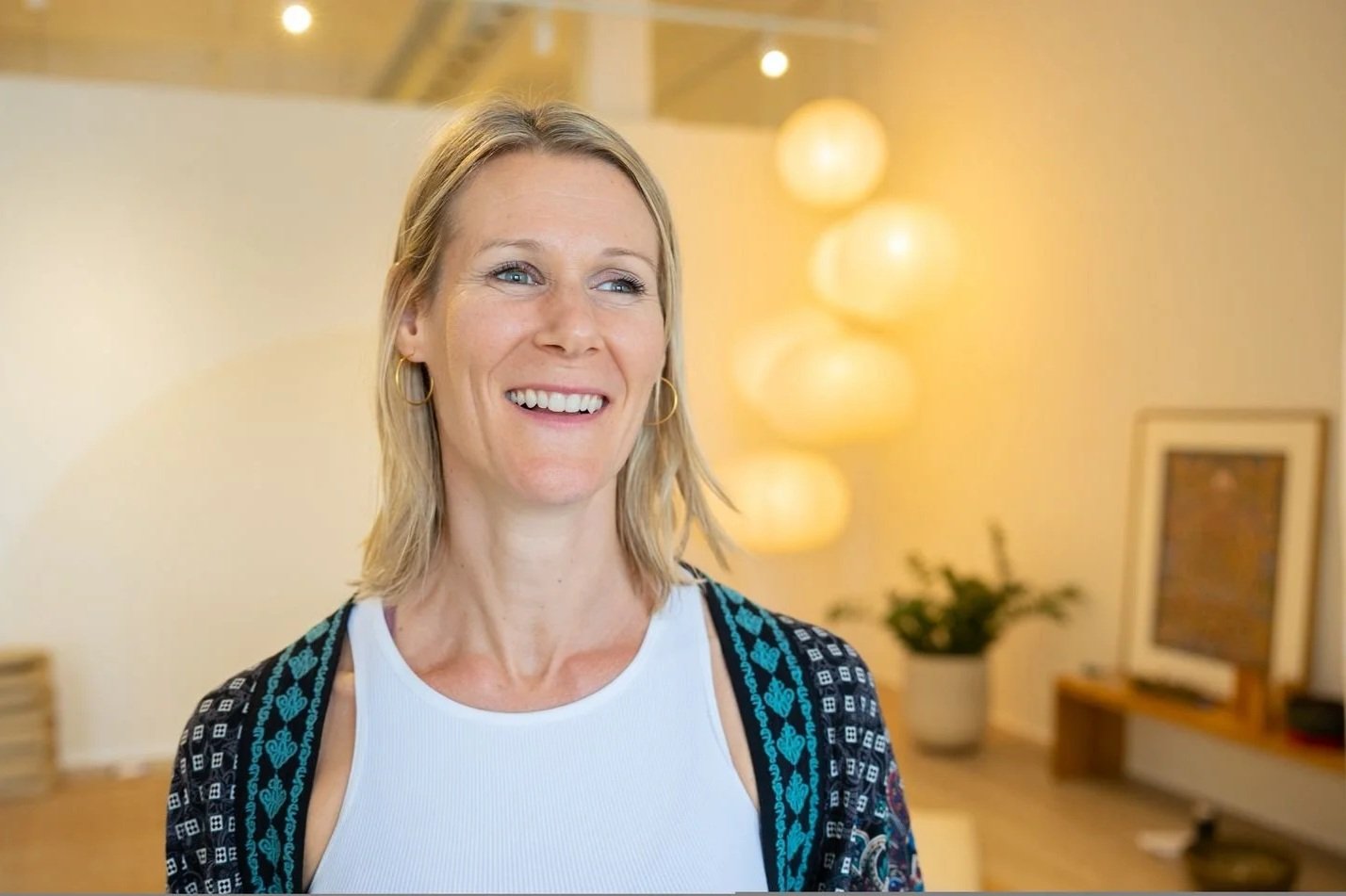Eva Still with blonde hair and a patterned cardigan, smiling in a brightly lit, modern living room with stylish hanging lamps.