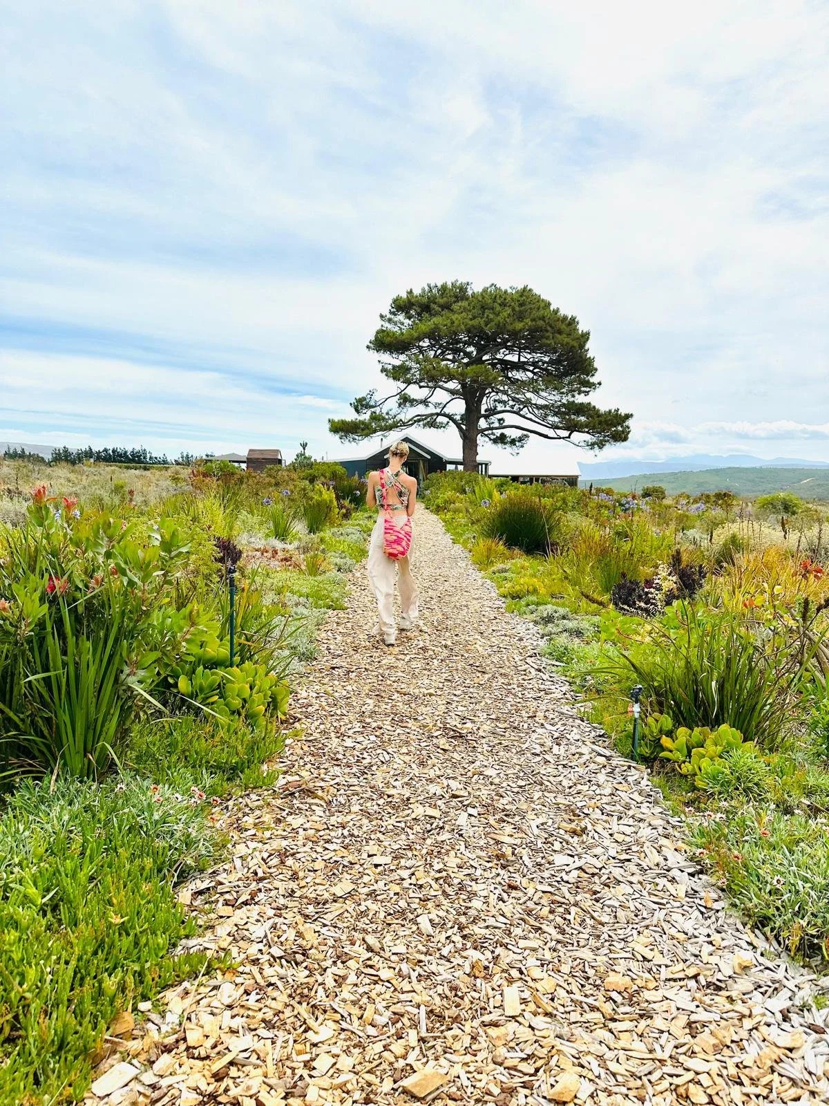 Eva Still walking along a gravel path through a lush garden with a large tree and a house in the background.