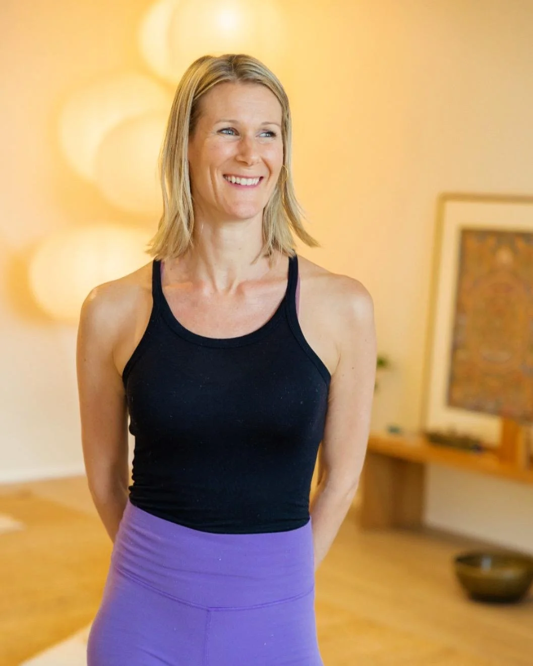 A woman with shoulder-length blonde hair wearing a black tank top and purple leggings, smiling in a warmly lit indoor space with blurred background decor.