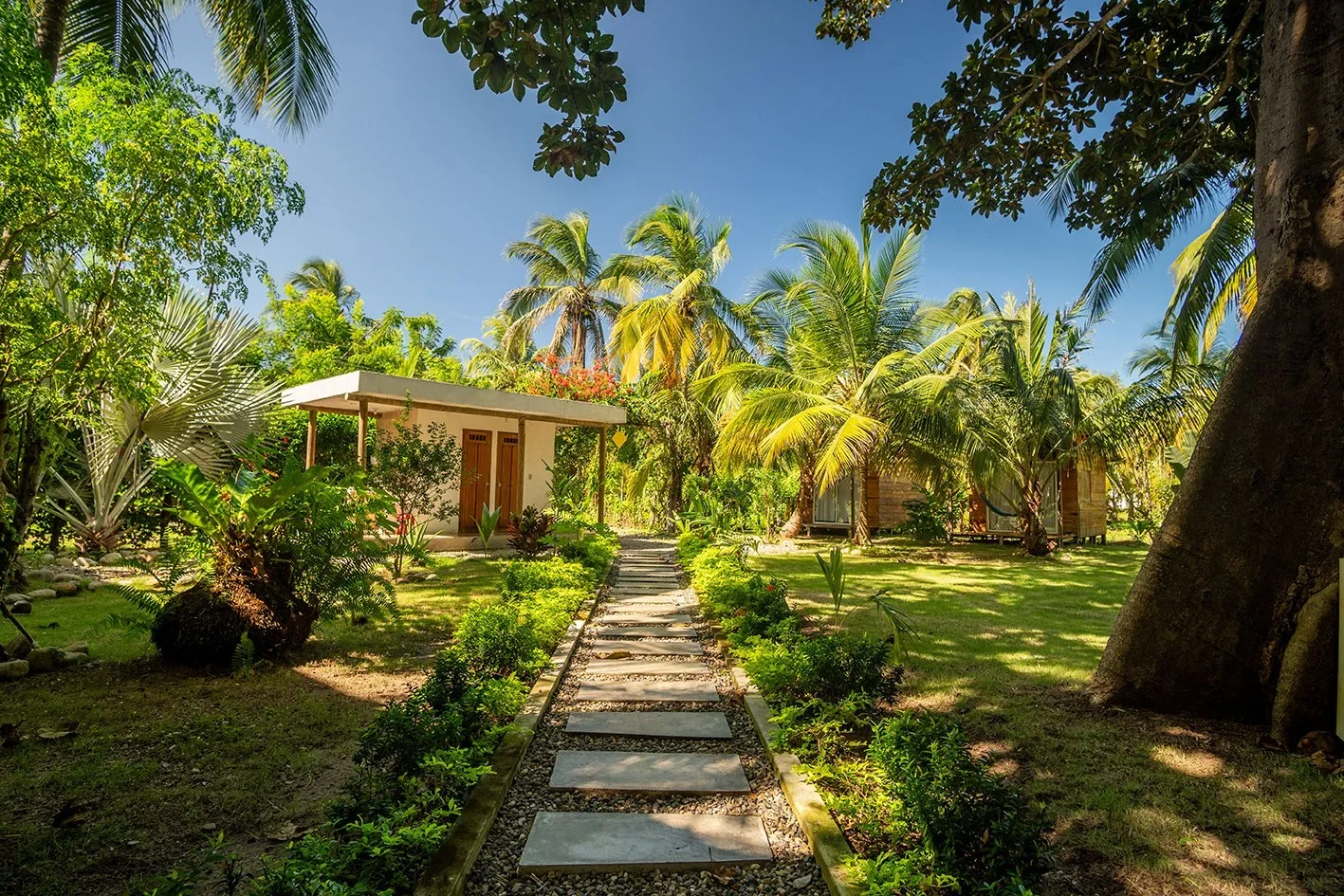 A garden with a stone pathway leading to small white and wooden cottages surrounded by lush green tropical plants and trees under a bright blue sky.
