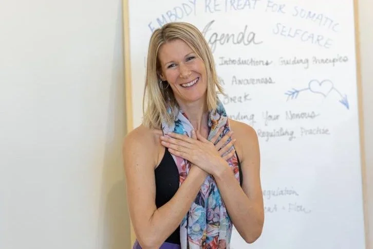 Eva Still wearing a colourful scarf and standing in front of a whiteboard featuring handwritten notes and a heart illustration.