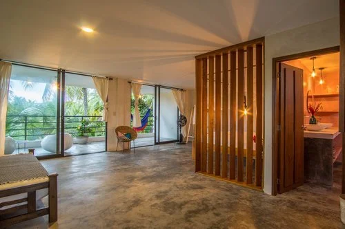 A spacious living area with large glass doors opening to a balcony with palm trees outside. The interior features a hammock, a chair, and a sitting area, with warm lighting and wooden accents.