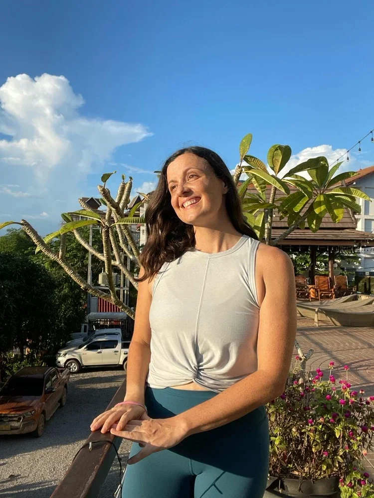 A woman smiling and looking away in an outdoor setting during the daytime, with a tree with large leaves, a building, parked cars, and a blue sky with clouds in the background.