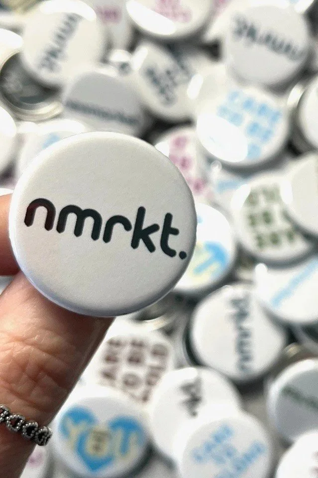 Close-up of a hand holding a white circular pin with the text 'nmrkt.' printed in black, with many similar pins in the background.