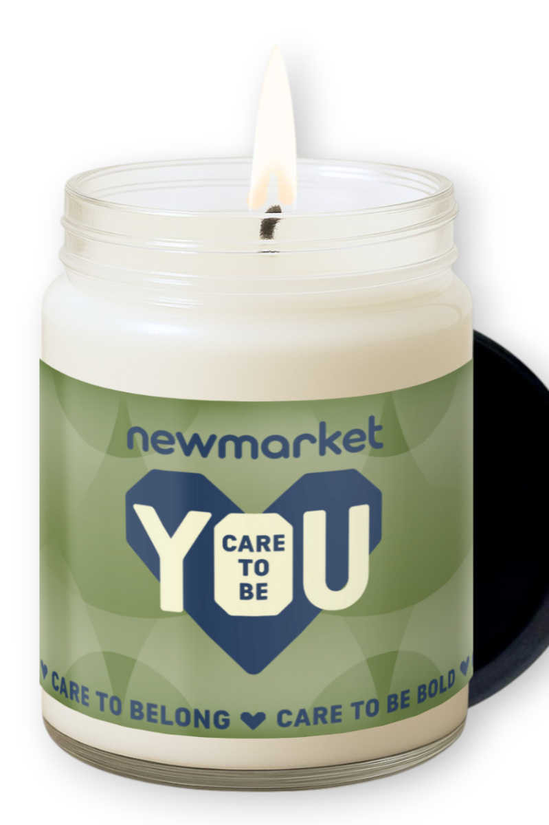 A candle in a glass jar with a lit wick, a black circle visible behind, and a green label that reads 'newmarket CARE TO BE YOU' with additional text 'CARE TO BELONG' and 'CARE TO BE BOLD'.