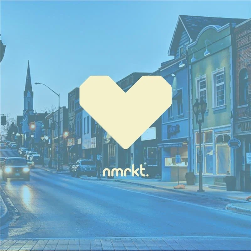 A street scene with buildings, cars, and a church steeple in the background, overlaid with a large cream-colored heart and the text 'nmrkt.' in the center.