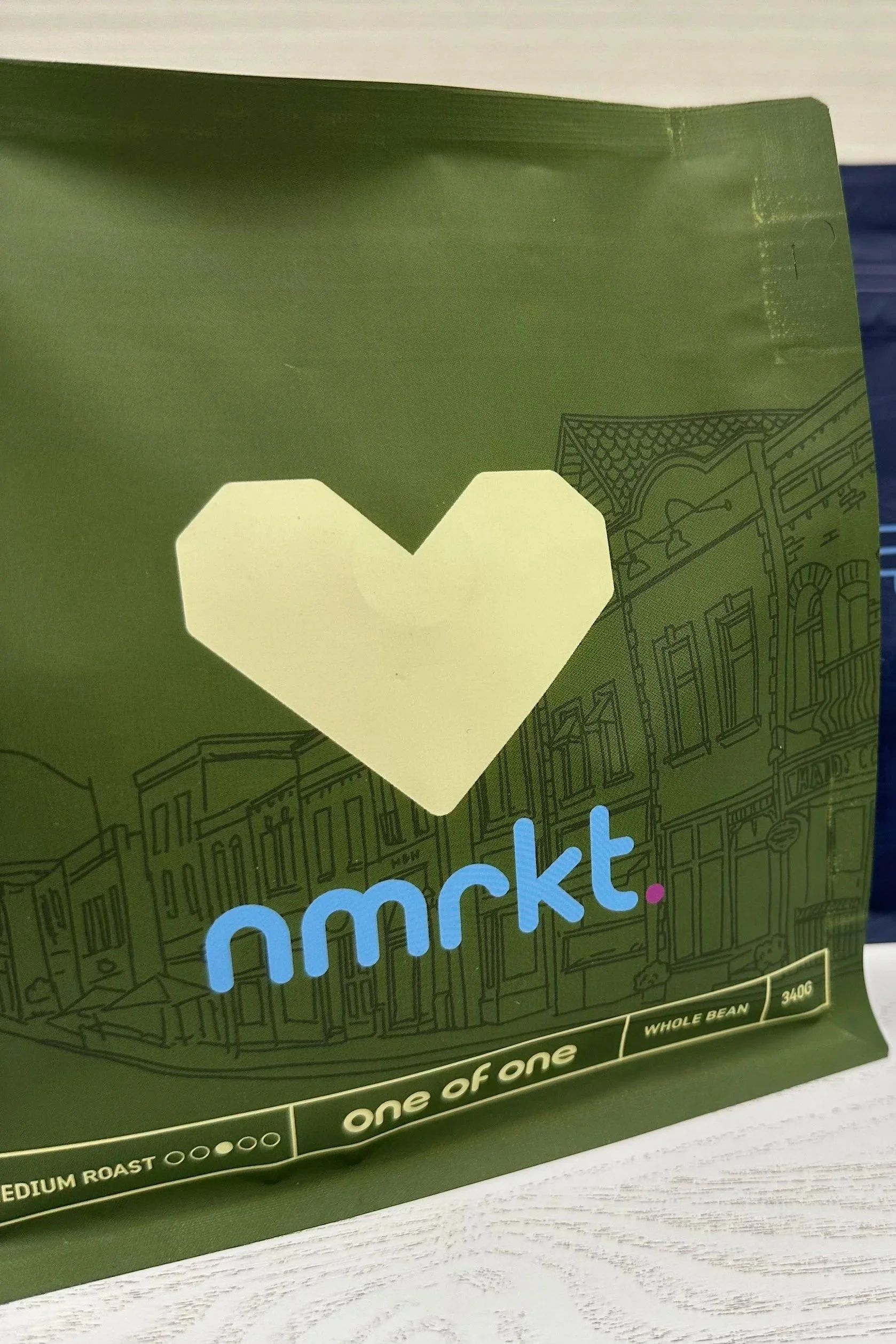 Close-up of a green coffee bean bag with a yellow heart-shaped label and the text 'nmrkt' in blue, along with information about medium roast and whole beans.