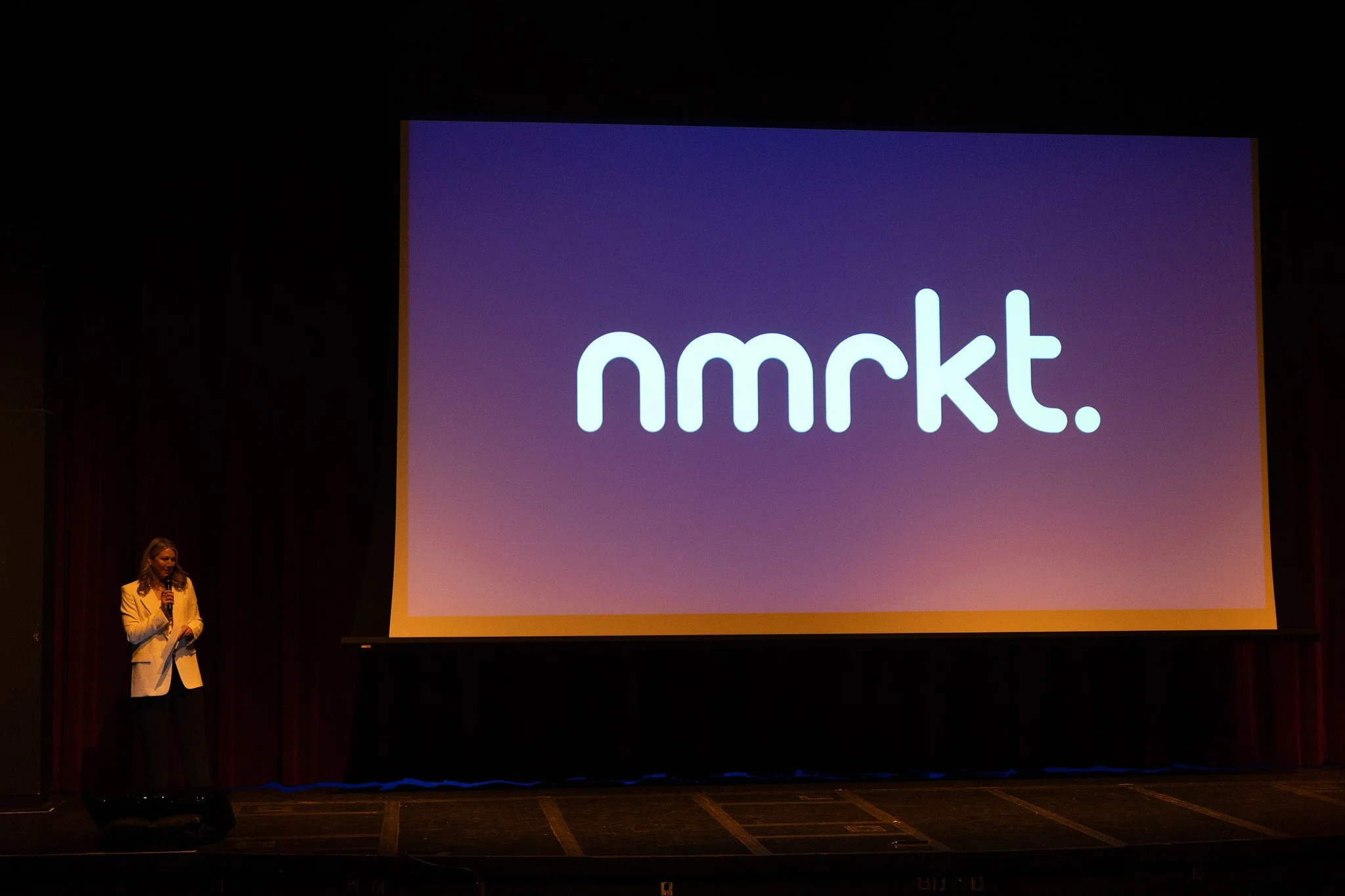 A woman in a beige blazer speaking into a microphone on a stage with a large screen displaying the logo 'nmrkt'.