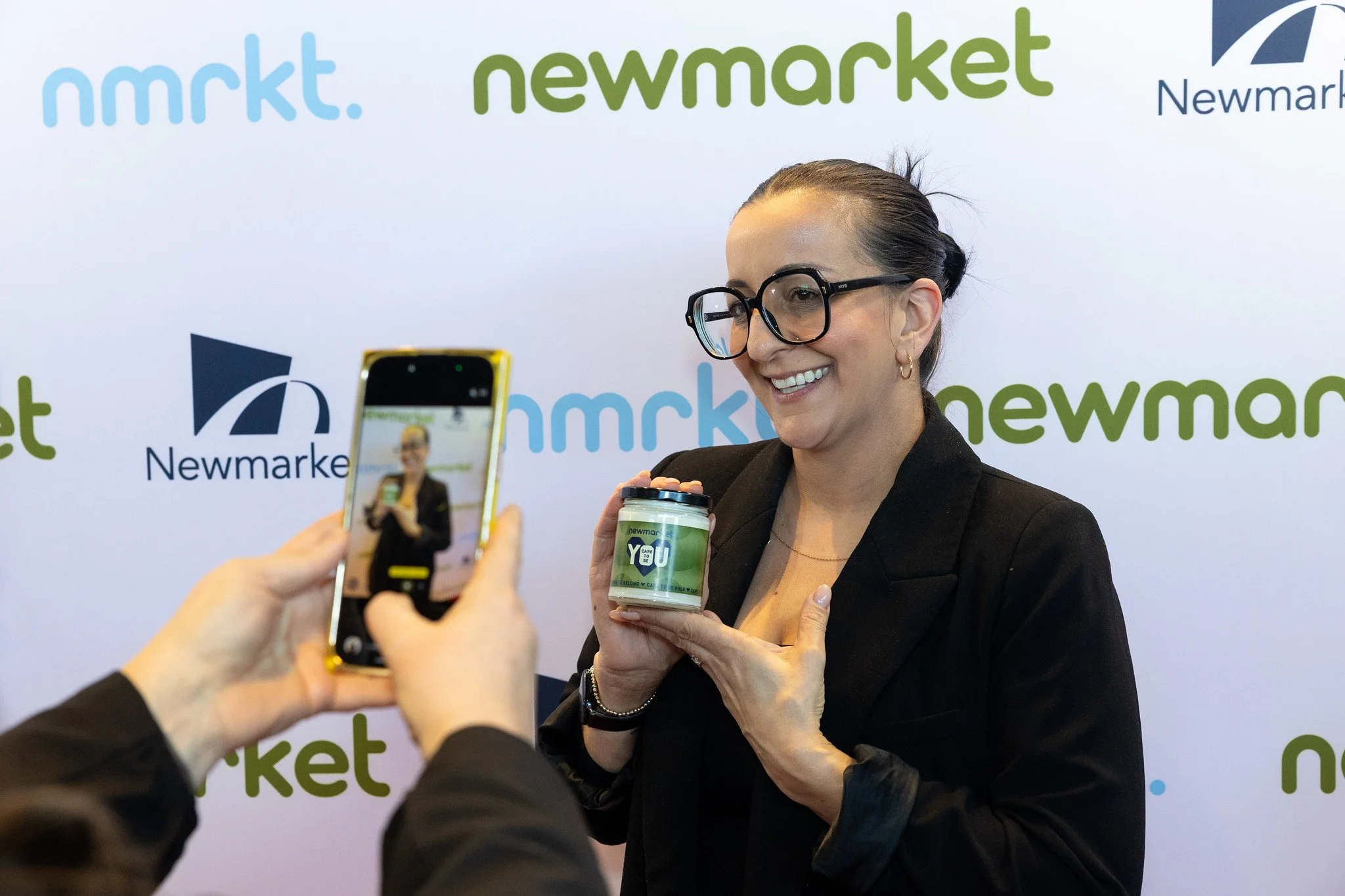A woman with glasses and earrings holding a container with a label that says "Care to Be You" and is smiling at a photo being taken of her, with a background displaying the words 'nmrkt. newmarket' and a logo.