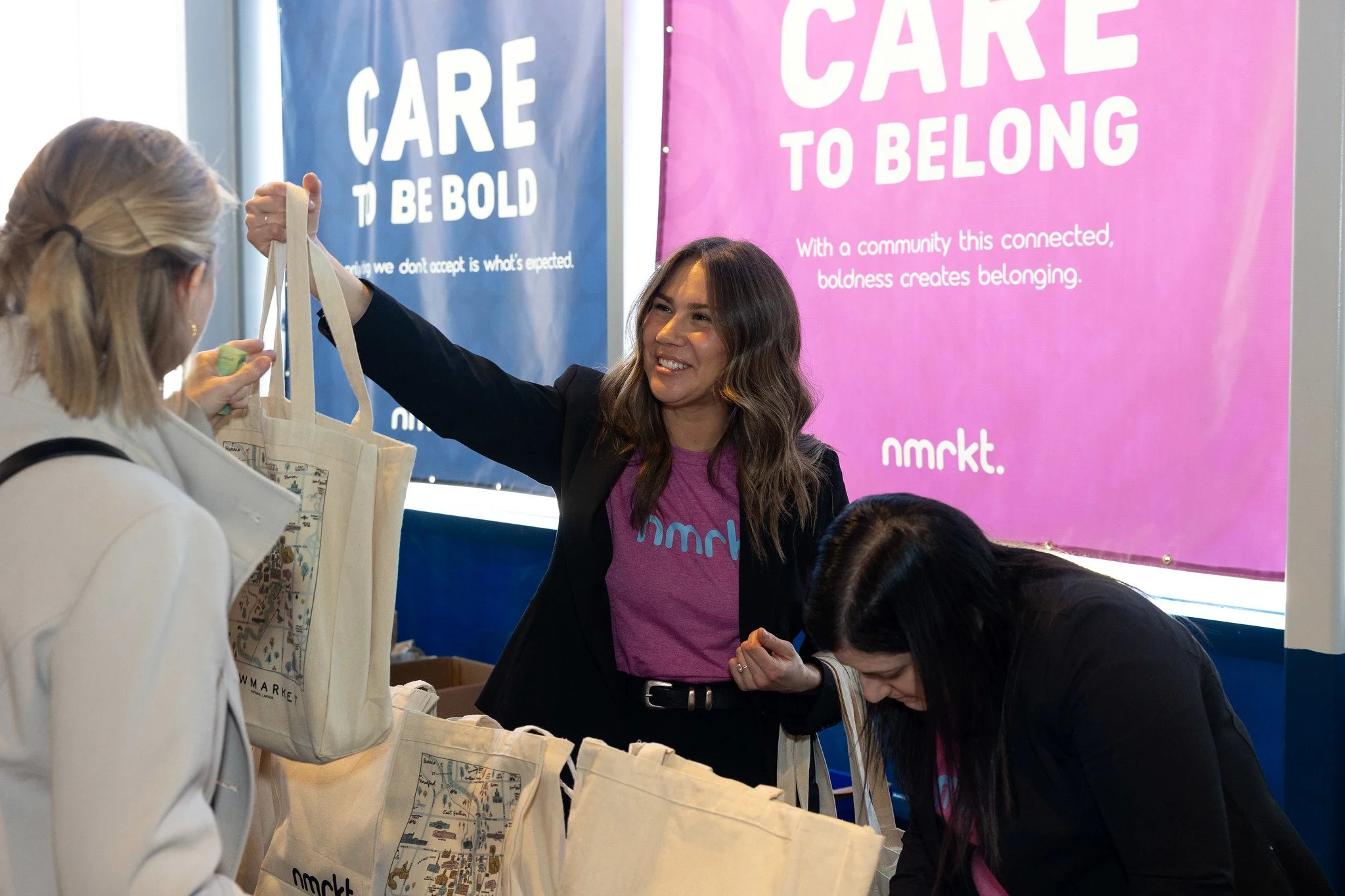 A woman smiling and handing a tote bag to another woman at an event with pink and blue banners in the background that read 'CARE TO BE BOLD' and 'CARE TO BELONG'.